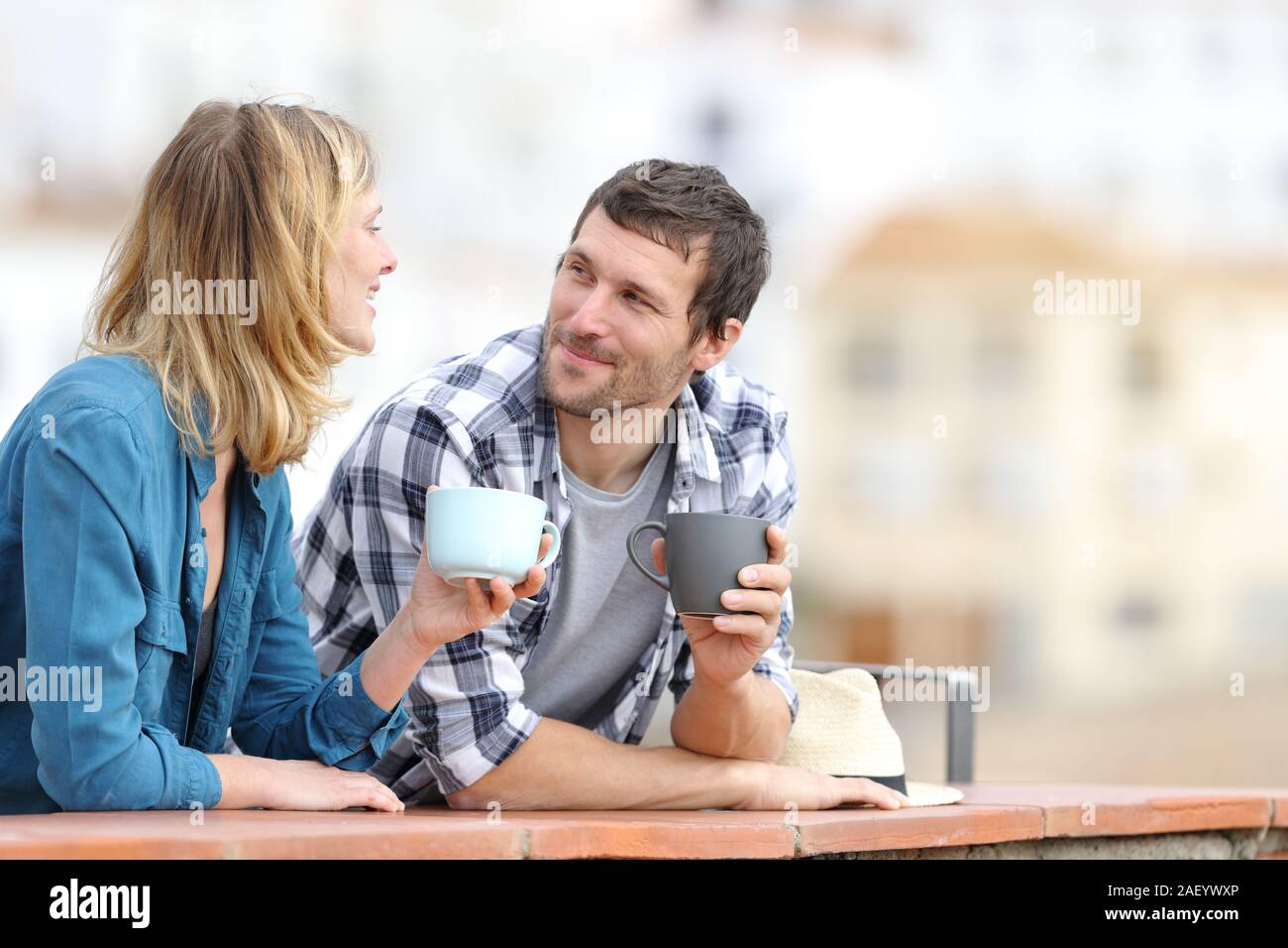Relaxed couple talking holding coffee mugs standing in a balcony outdoors in a rural town Stock Photo