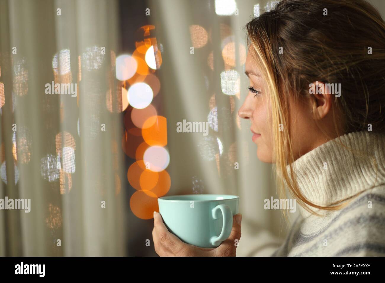 Woman holding coffee cup standing looking at city lights through a window in the night at home Stock Photo