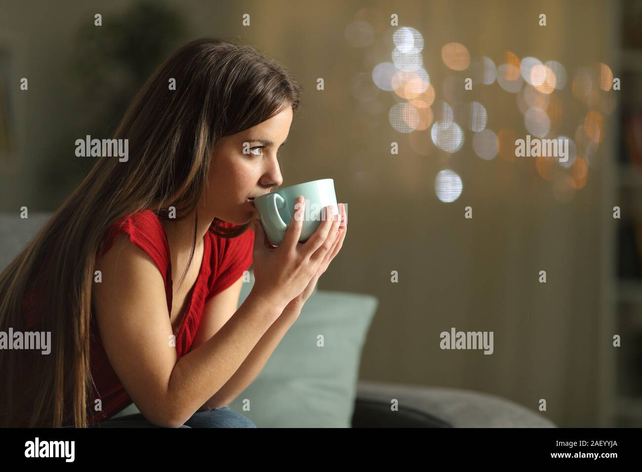 Side view portrait of a woman looking away drinking coffee sitting on a couch in the night at home Stock Photo