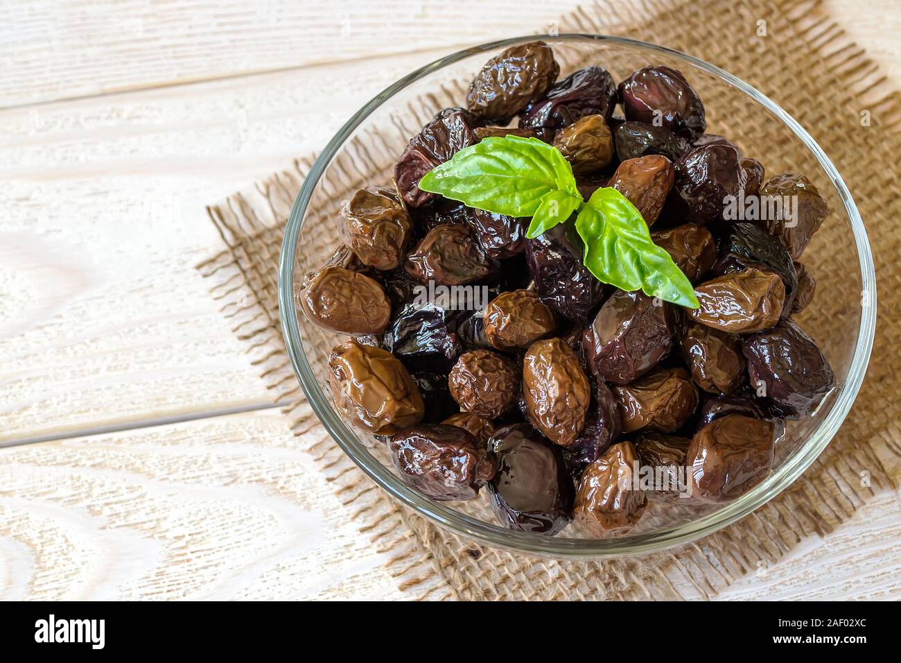 Sun-dried or cured olives in a glass bowl on a white rustic wooden table. Healhy eating and vegetarian food. Ingredient for tapenade, sandwich, salad. Stock Photo
