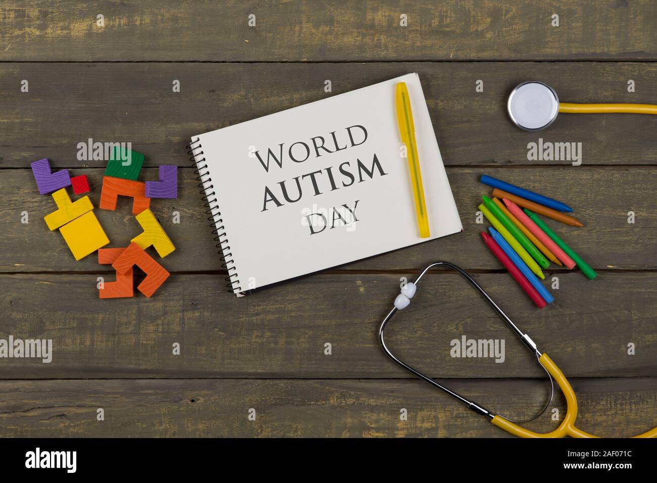 Mental health concept - text World Autism day, notepad, yellow stethoscope, colorful wooden jigsaw puzzles, crayons on wooden background Stock Photo
