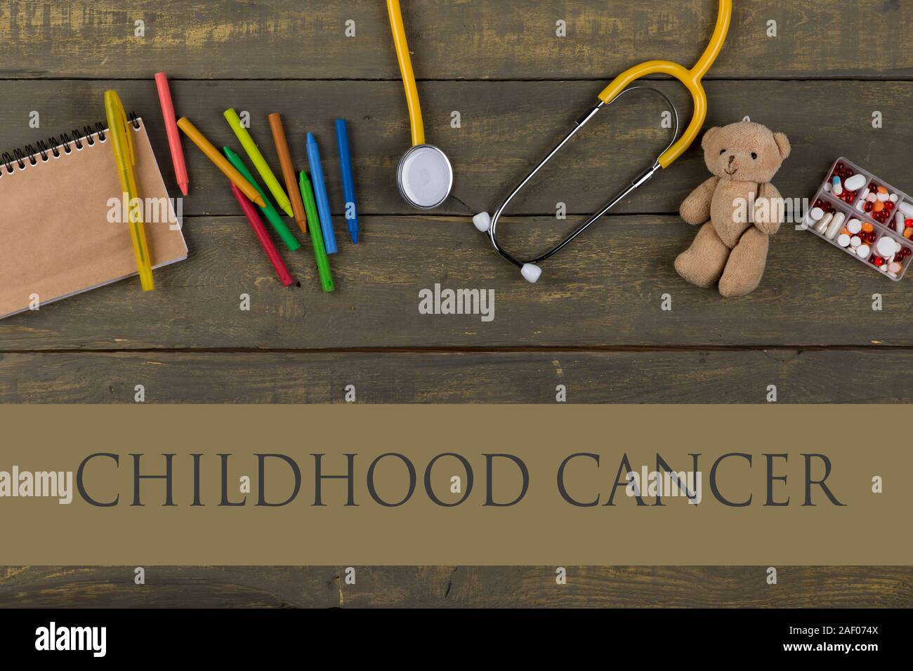 dangerous disease prevention concept - text Childhood cancer, notepad, pills, yellow stethoscope, Teddy bear toy, crayons on wooden background. Top vi Stock Photo