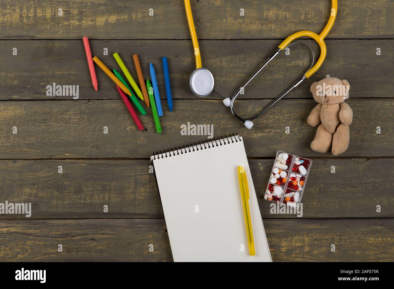 Children's doctor or pediatrician concept - blank notepad, pills, yellow stethoscope, Teddy bear toy, crayons on wooden background. Top view Stock Photo