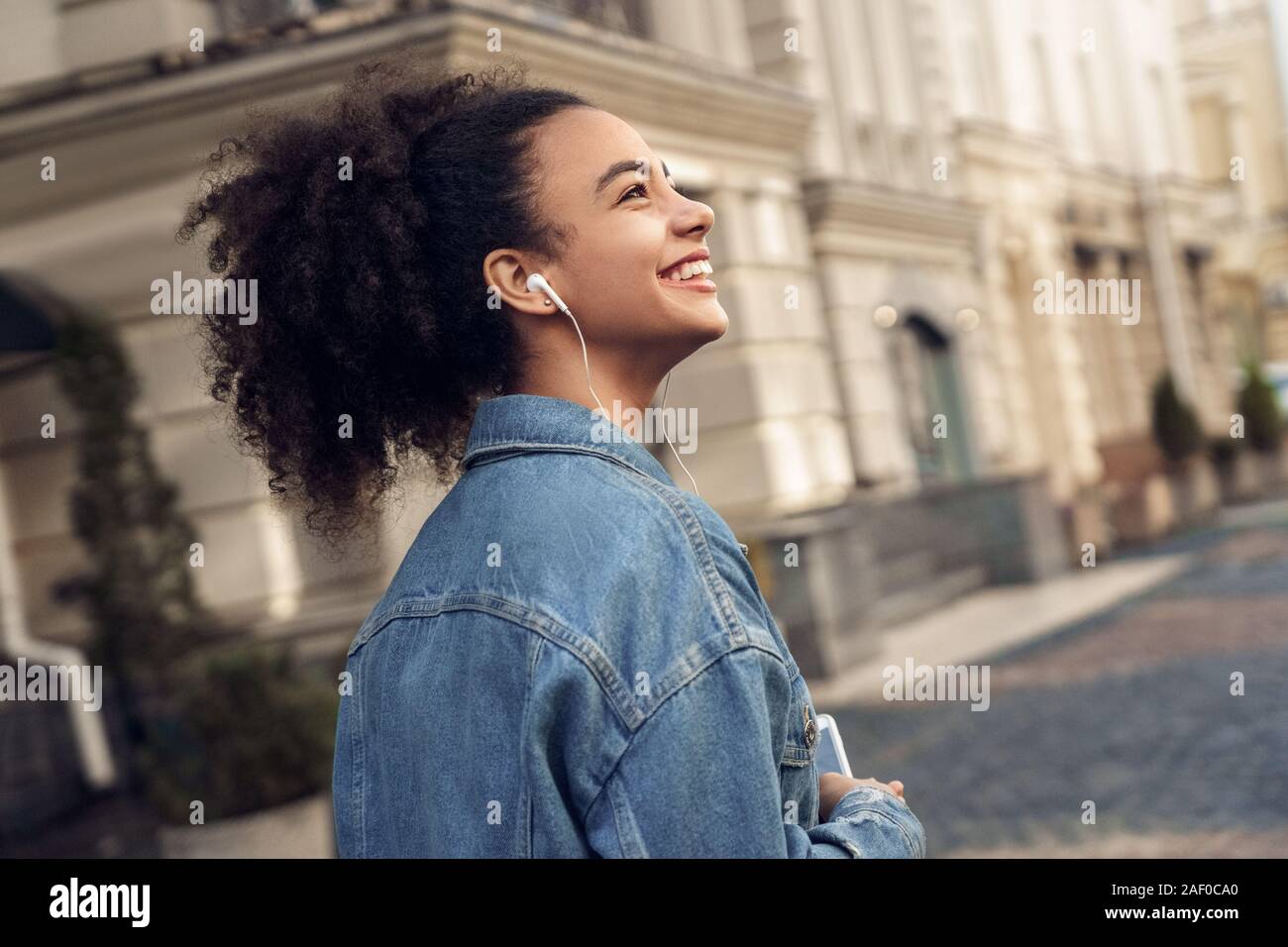 Outdoors Leisure. Young stylish african girl in denim jacket and earphones standing listening music looking aside cheerful back view Stock Photo Alamy