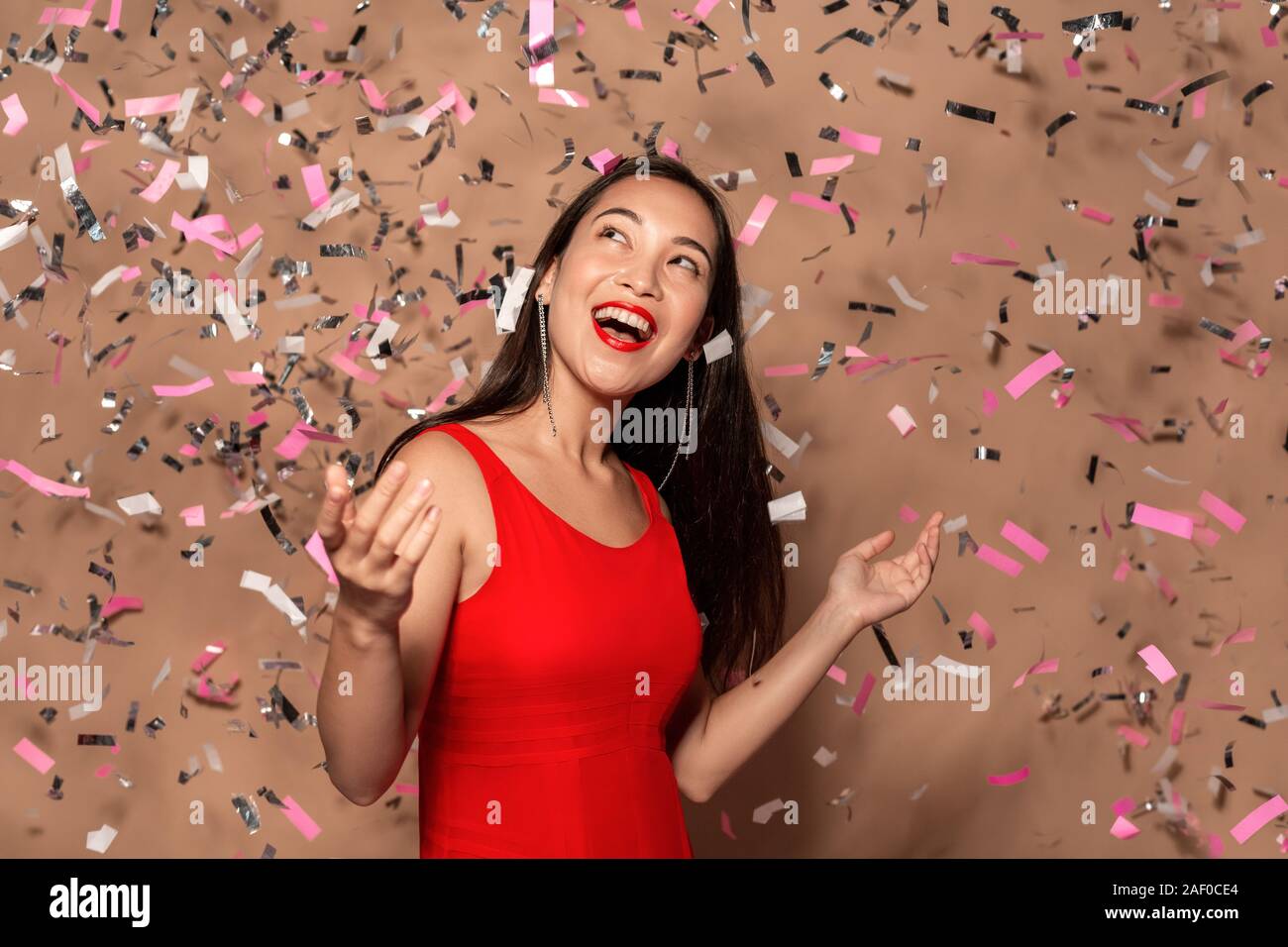 Freestyle. Young girl in dress standing in falling confetti isolated on brown looking up smiling surprised Stock Photo