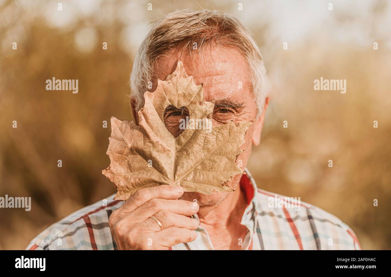 The senior man looking through the heart-shaped hole in the maple leaf. Autumn concept. Selective focus Stock Photo