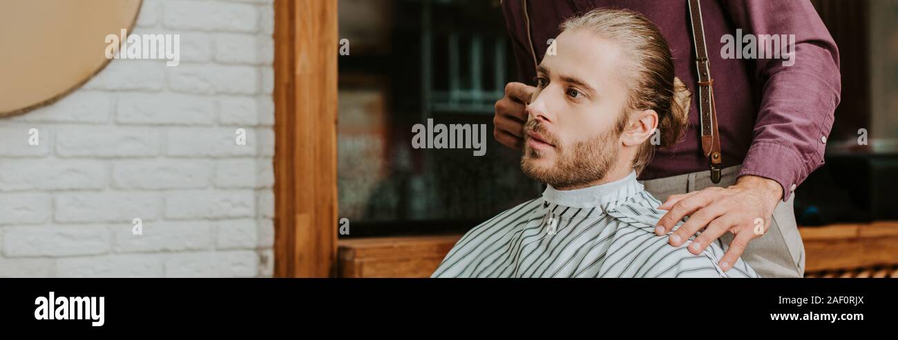 panoramic shot of barber touching hair of handsome man in barbershop Stock Photo