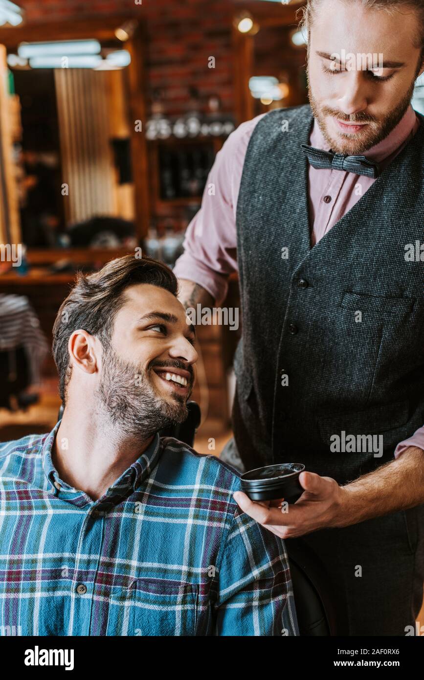handsome barber holding jar with black hair pomade near happy man Stock Photo