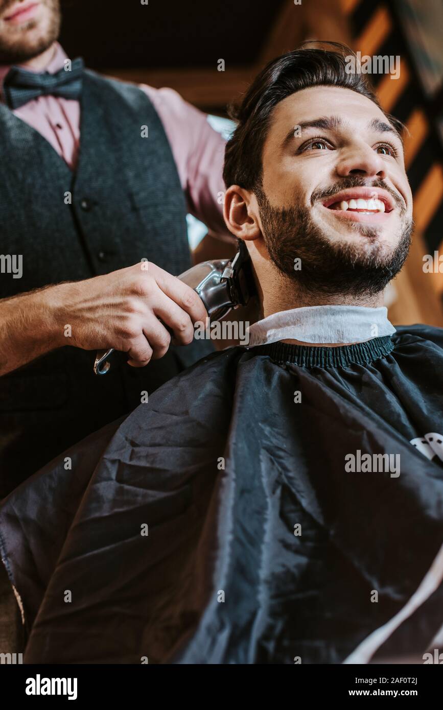 low angle view of barber holding trimmer while styling hair of cheerful man in barbershop Stock Photo