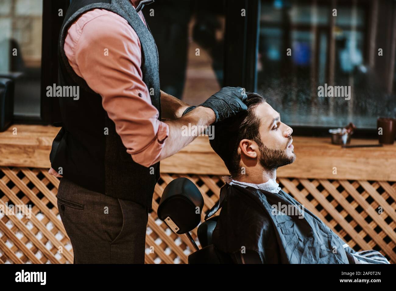 side view of bearded man sitting near barber in black latex gloves Stock Photo