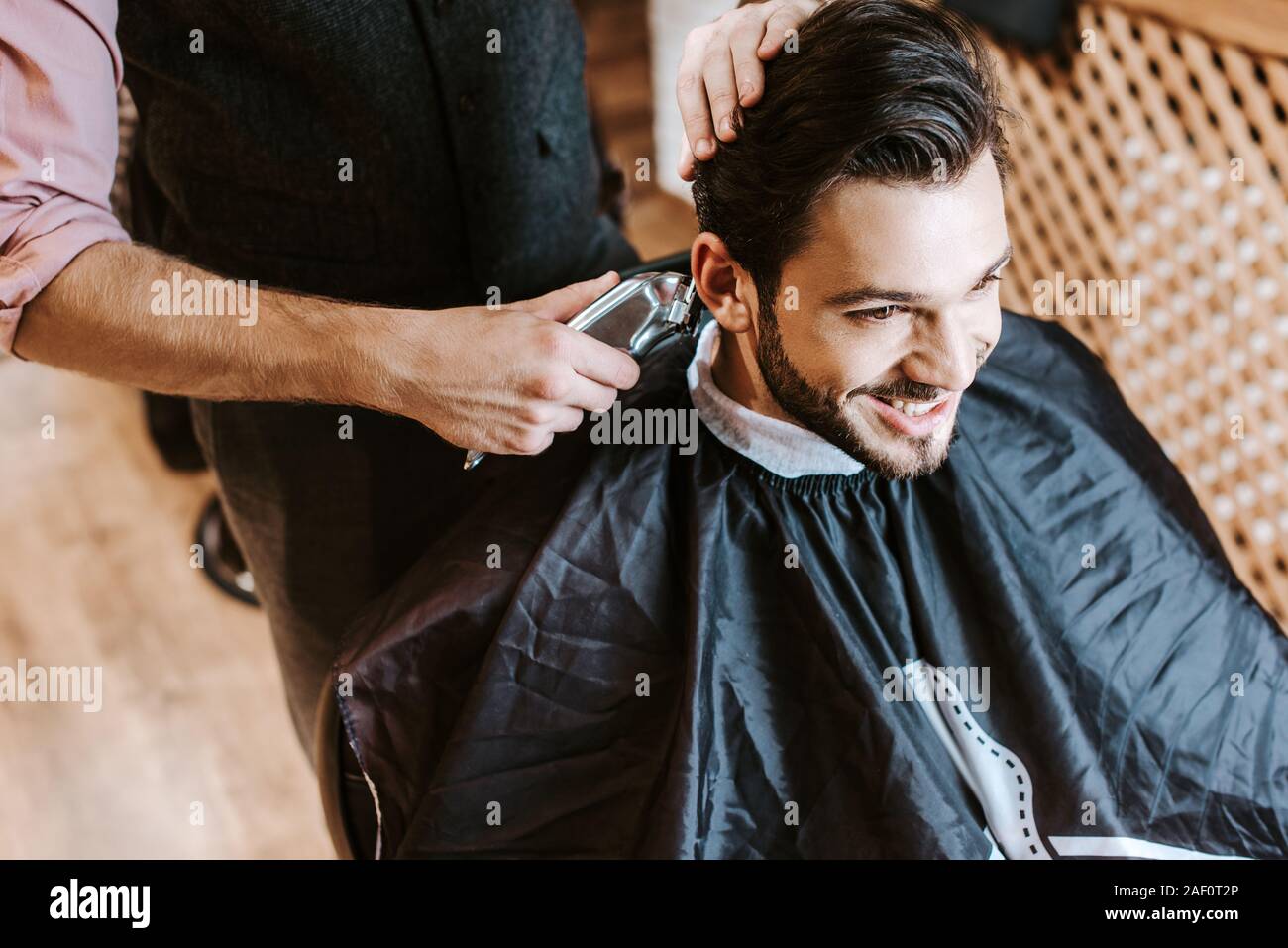 barber holding trimmer while styling hair of happy man in barbershop Stock Photo
