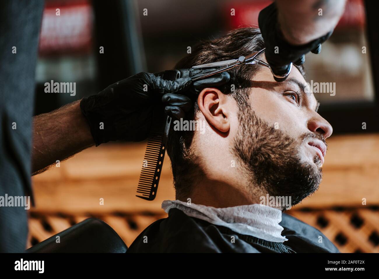 selective focus of barber in latex gloves holding hair comb and scissors while styling hair of man Stock Photo