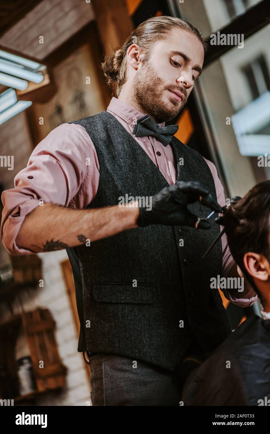 tattooed barber in latex gloves holding hair comb and scissors while styling hair of man Stock Photo