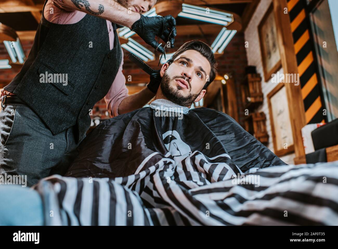 low angle view of tattooed barber in latex gloves holding scissors while styling hair of man Stock Photo