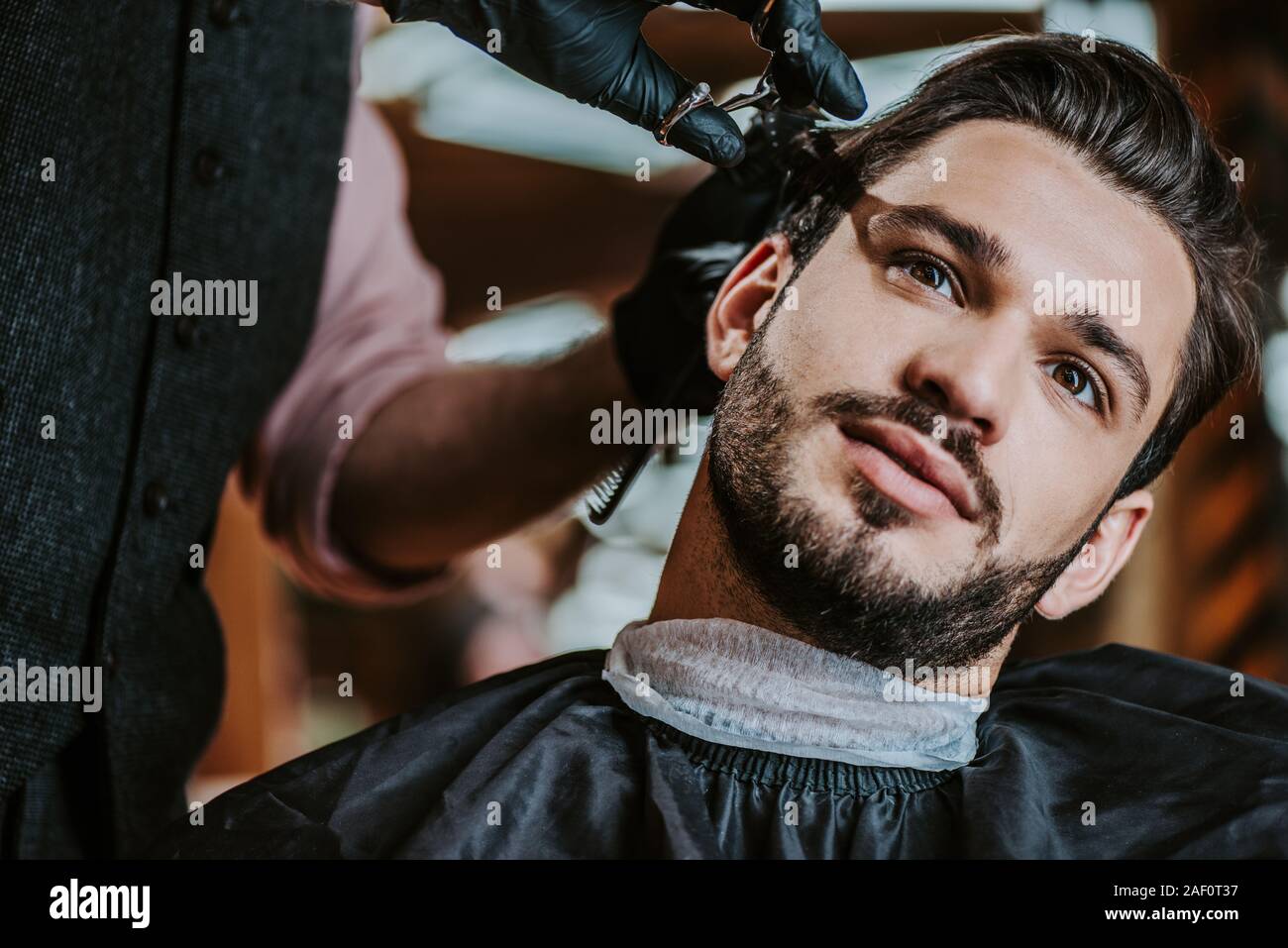 selective focus of barber in latex gloves holding scissors while styling hair of man Stock Photo