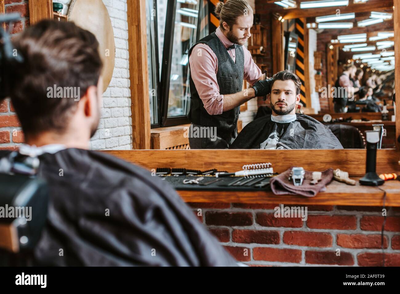 selective focus of handsome barber styling hair of man near mirror in barbershop Stock Photo