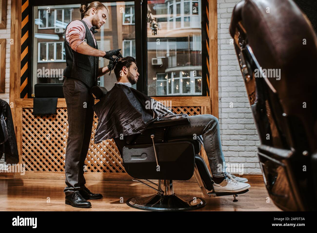 selective focus of handsome barber styling hair of man in modern barbershop Stock Photo