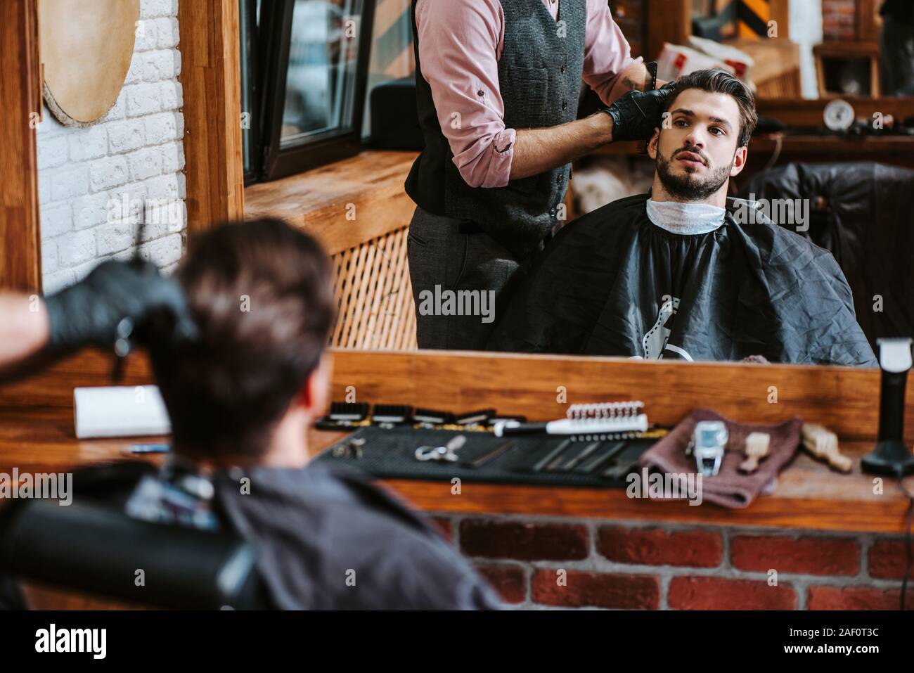 selective focus of barber styling hair of man near mirror in barbershop Stock Photo