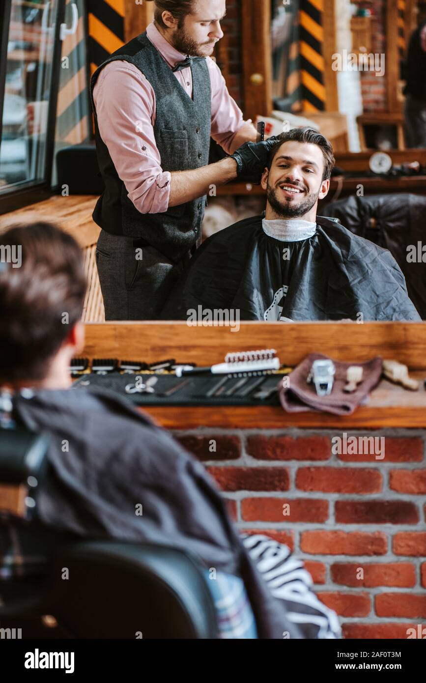 selective focus of handsome barber styling hair of happy man near mirror in barbershop Stock Photo