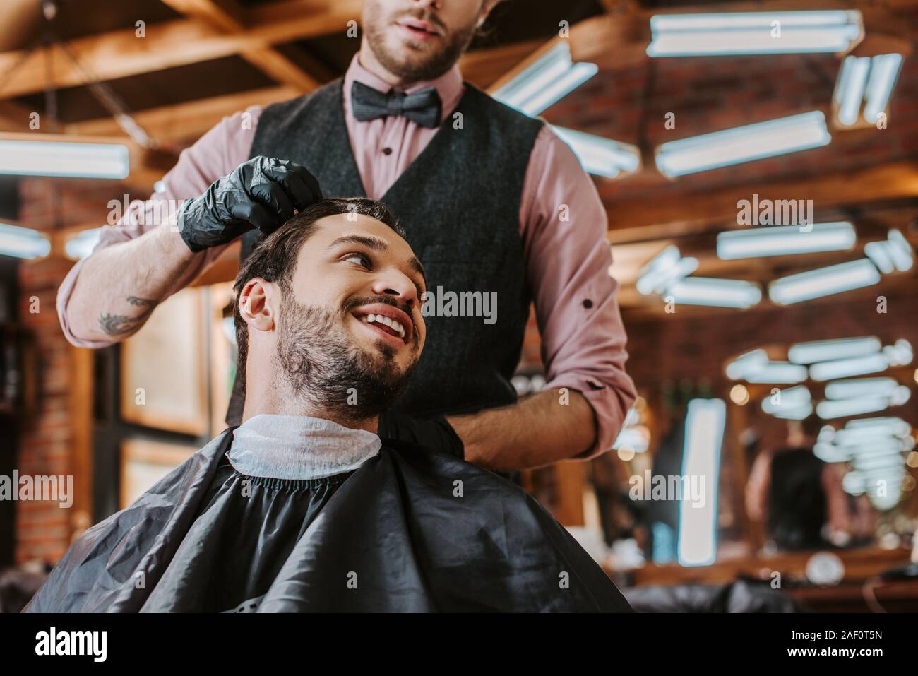 bearded barber in black latex gloves touching hair of happy man in barbershop Stock Photo