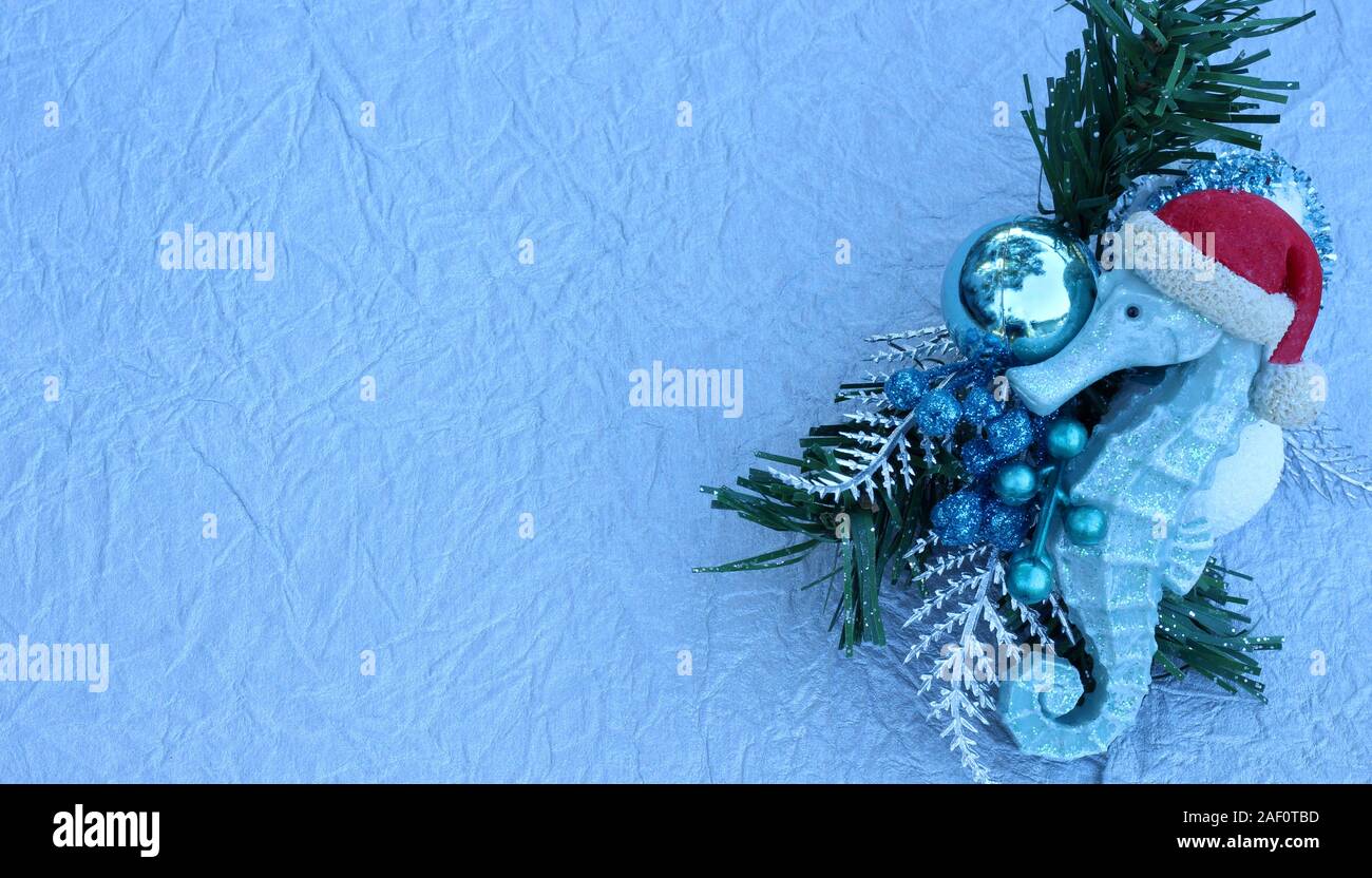 turquoise seahorse with red Santa Claus hat laying on a pine branch with blue christmas bulbs on festive silver background with copy space Stock Photo