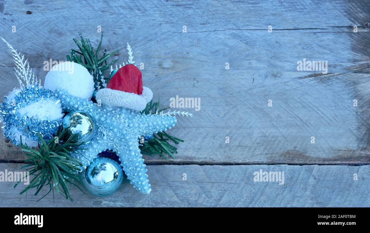 turquoise starfish with red Santa Claus hat laying on a pine branch with christmas bulbs on a natural wood background with copy space Stock Photo