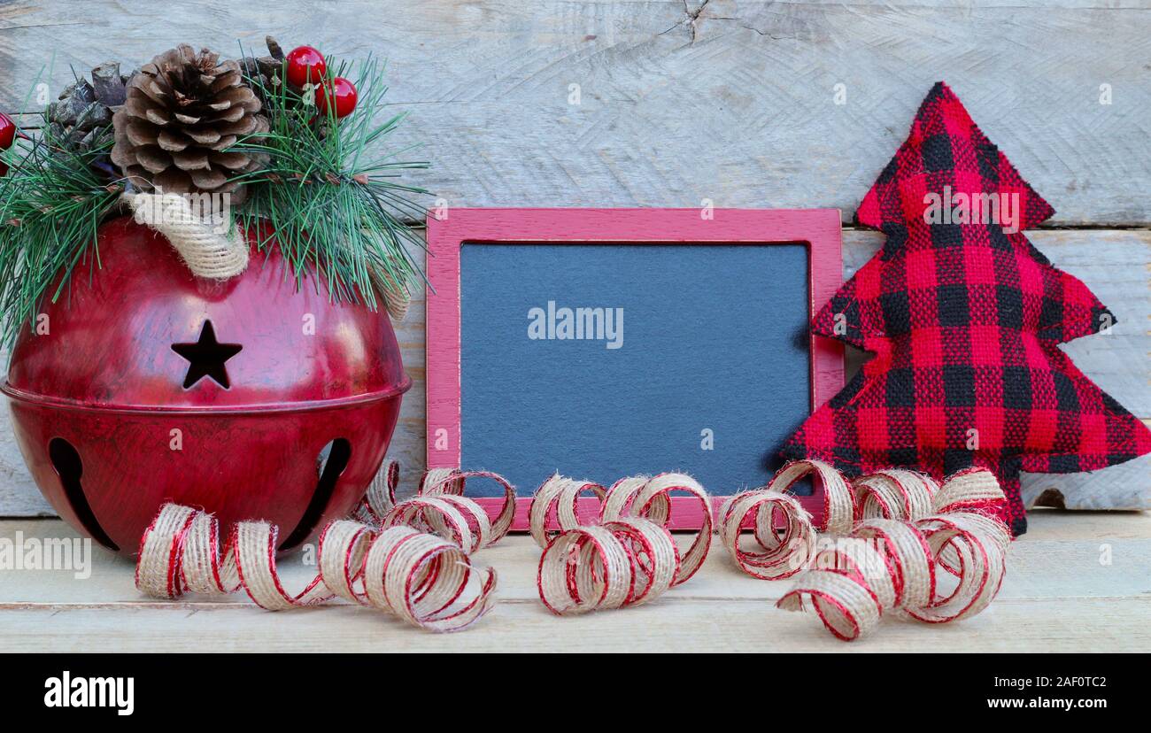 red jingle bell with pine and berries next to a red framed chalkboard, cloth christmas tree and curled ribbon on a wood background with copy space Stock Photo