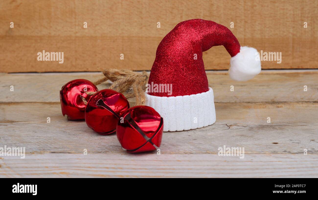 red and white Santa hat next to three jingle bells on a wooden background with writing space Stock Photo