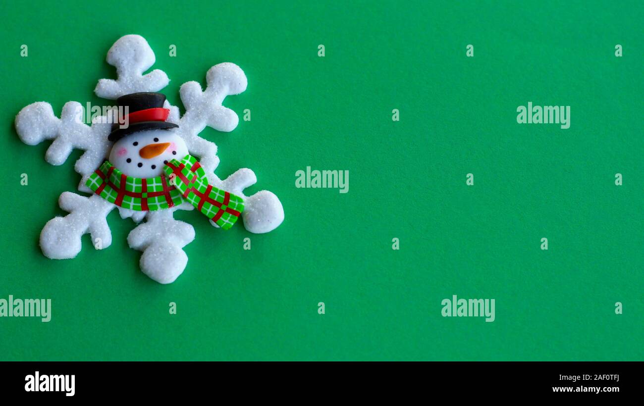 white snowflake decorated with a snowman face in the center wearing a hat and scarf on a green background with copy space Stock Photo