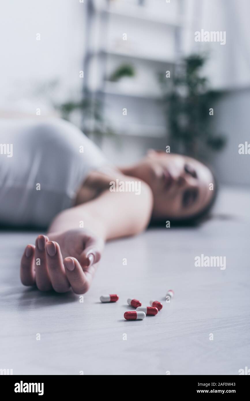 selective focus of lifeless girl, committed suicide by overdosing medicines, lying on floor near scattered pills Stock Photo