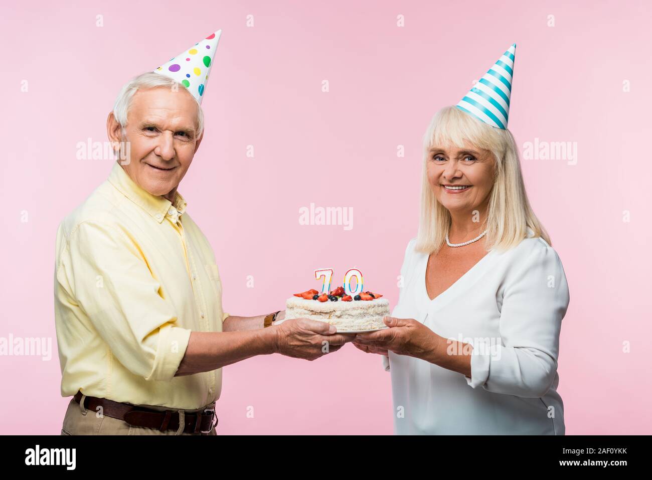 happy senior couple in party caps holding birthday cake isolated on pink Stock Photo