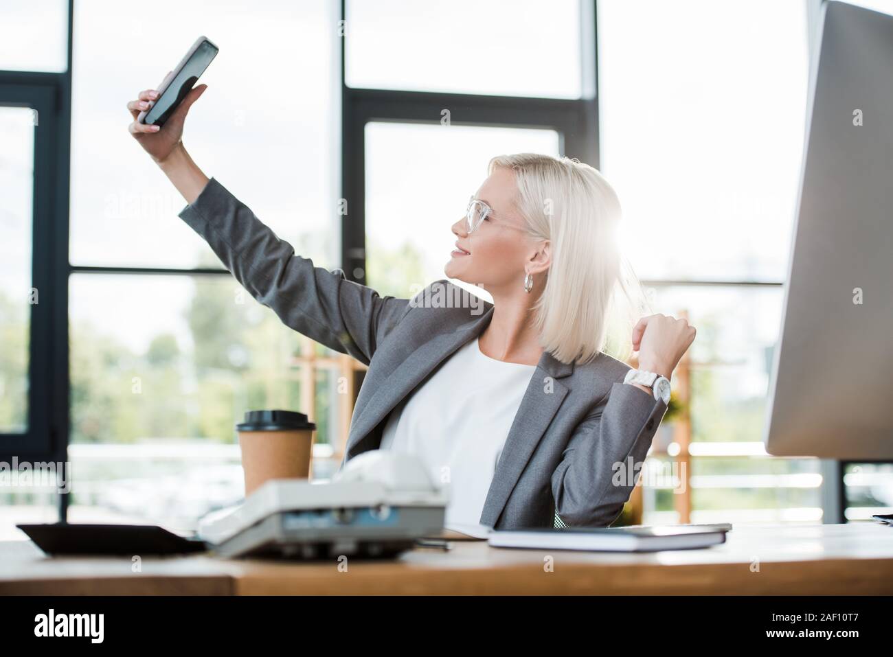 selective focus of cheerful businesswoman in glasses taking selfie in modern office Stock Photo
