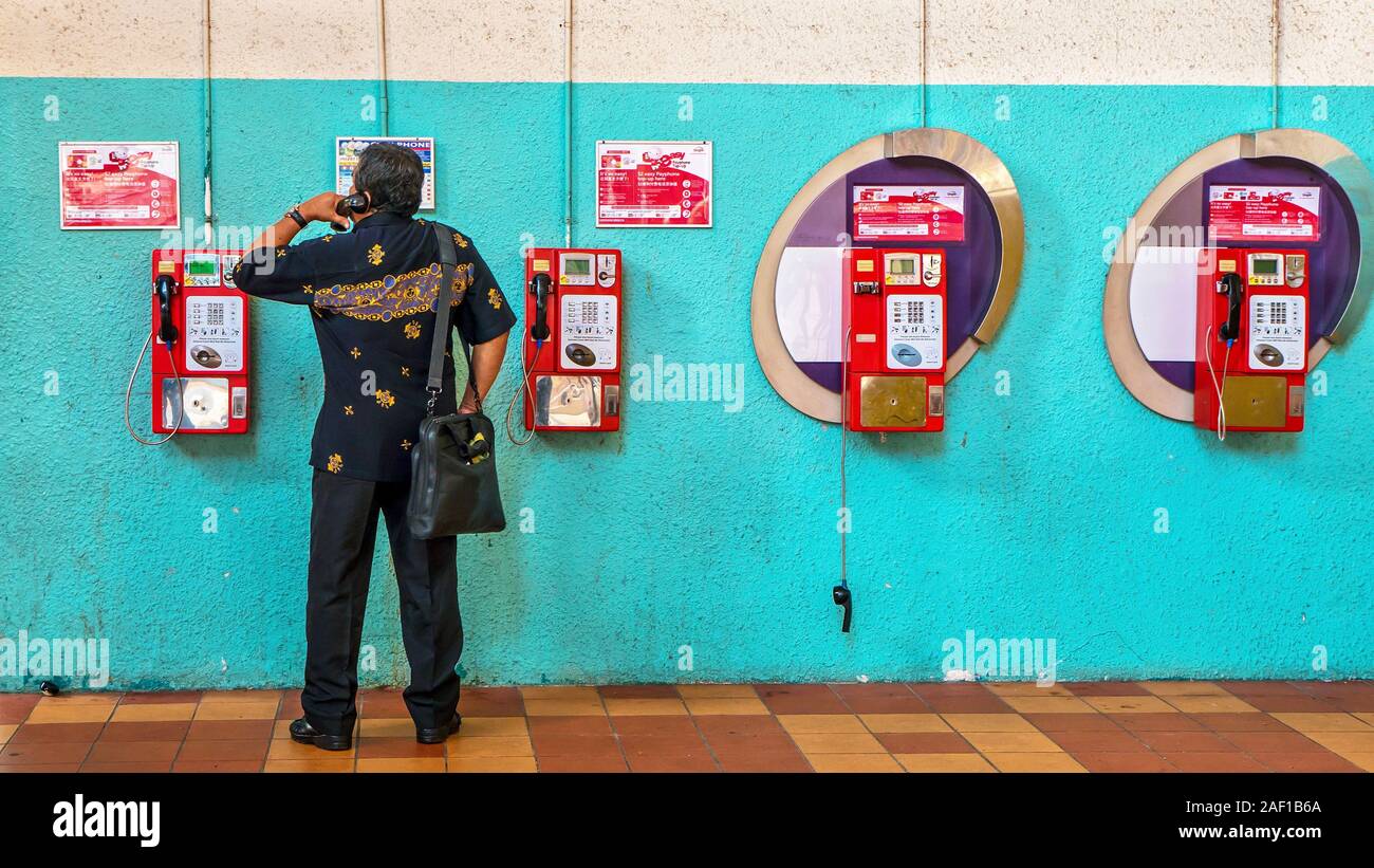 An Asian man using the old-fashioned, coin-operated, push button, wall-mounted payphone system, with plastic handsets in Singapore. Stock Photo