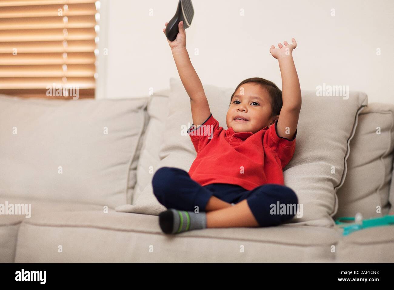 A happy kid with arms raised up while watching tv in the living room and sitting on the couch. Stock Photo