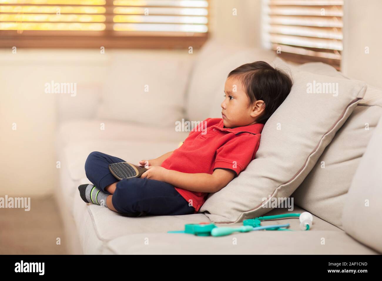 A little boy comfortably sitting on a living room couch and holding a remote control while watching tv. Stock Photo