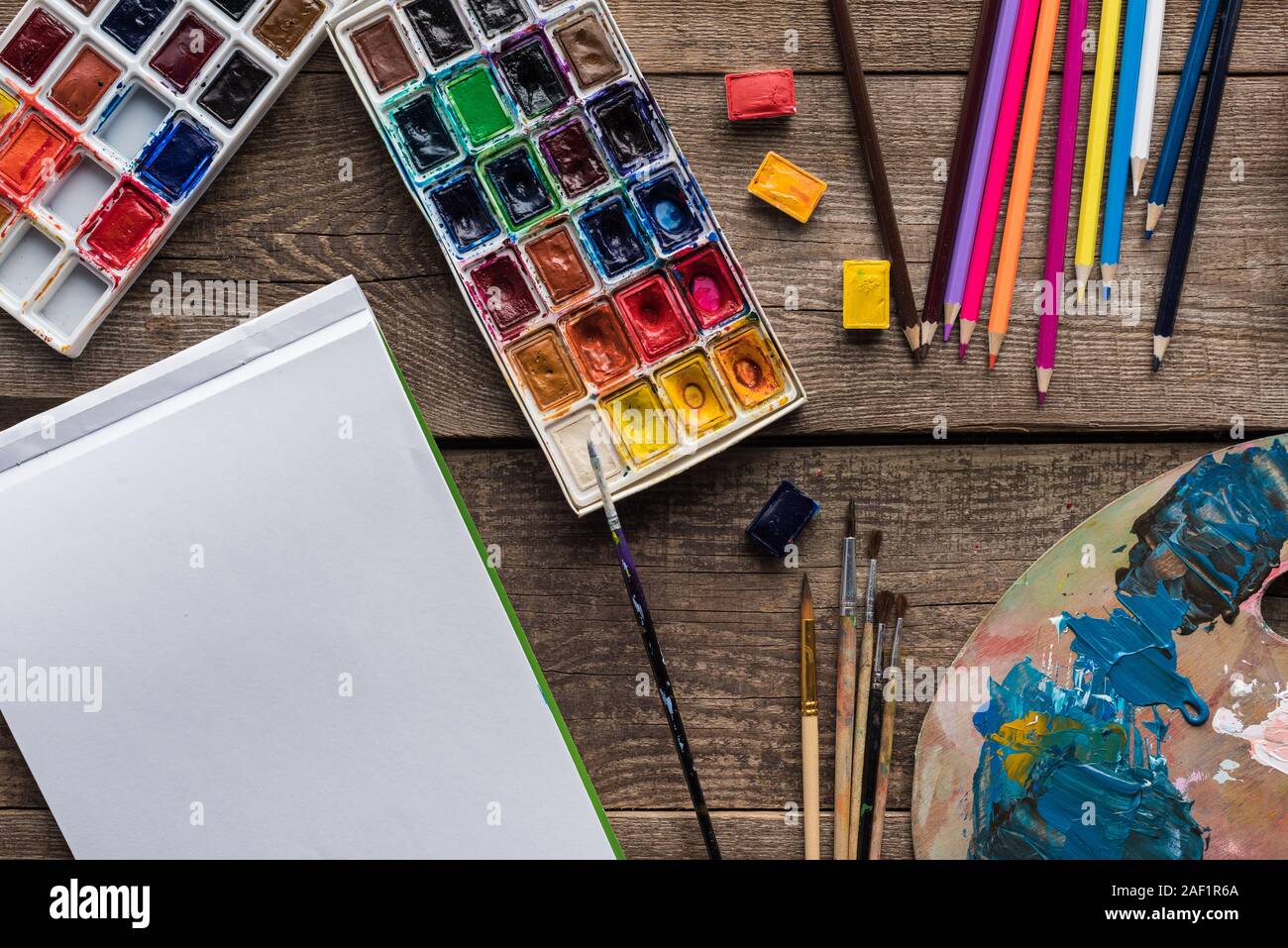 https://c8.alamy.com/comp/2AF1R6A/top-view-of-paint-palettes-paintbrushes-color-pencils-and-blank-sketch-pad-on-wooden-surface-2AF1R6A.jpg