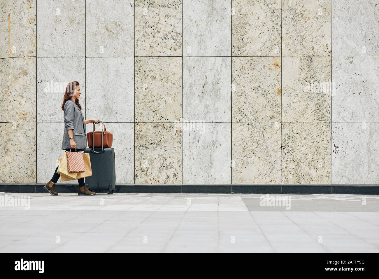 Positive Vietnamese woman with big suitcase and paperbags walking to