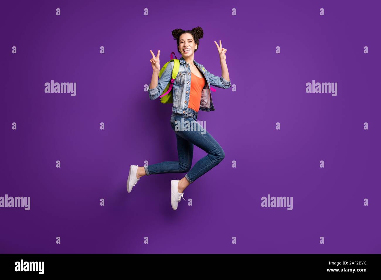 Full length profile photo of funny student lady jumping high showing v-sign symbols wear green bag casual denim stylish outfit isolated purple color Stock Photo