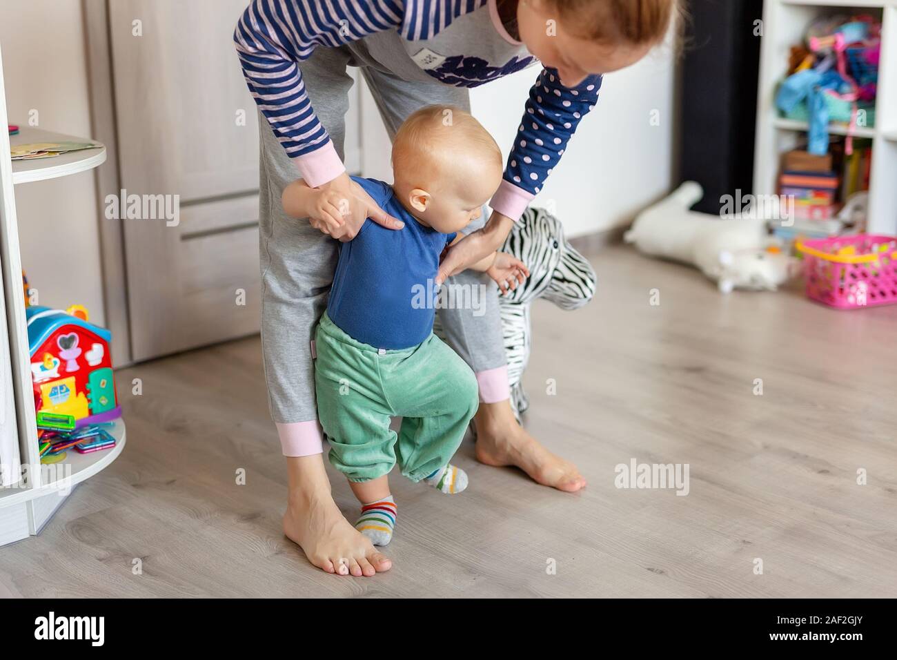 Cute little adorable blond toddler boy making first steps with mother support in playroom at home. Happy funny child learning to walk with mom help Stock Photo