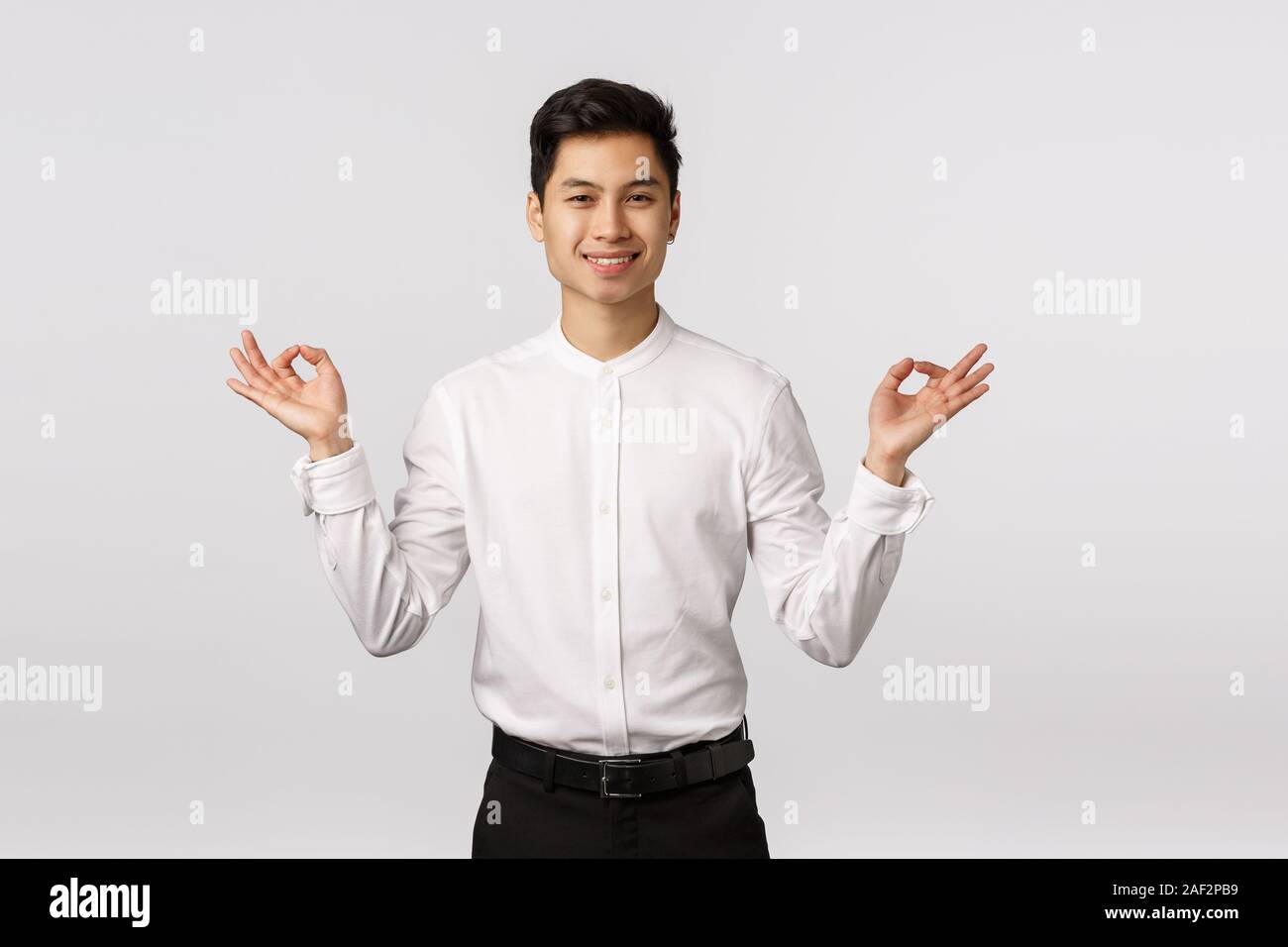 Peaceful and carefree, smiling relaxed cute asian businessman in white shirt, pants, formal outfit, trying take rest from business worries, meditating Stock Photo