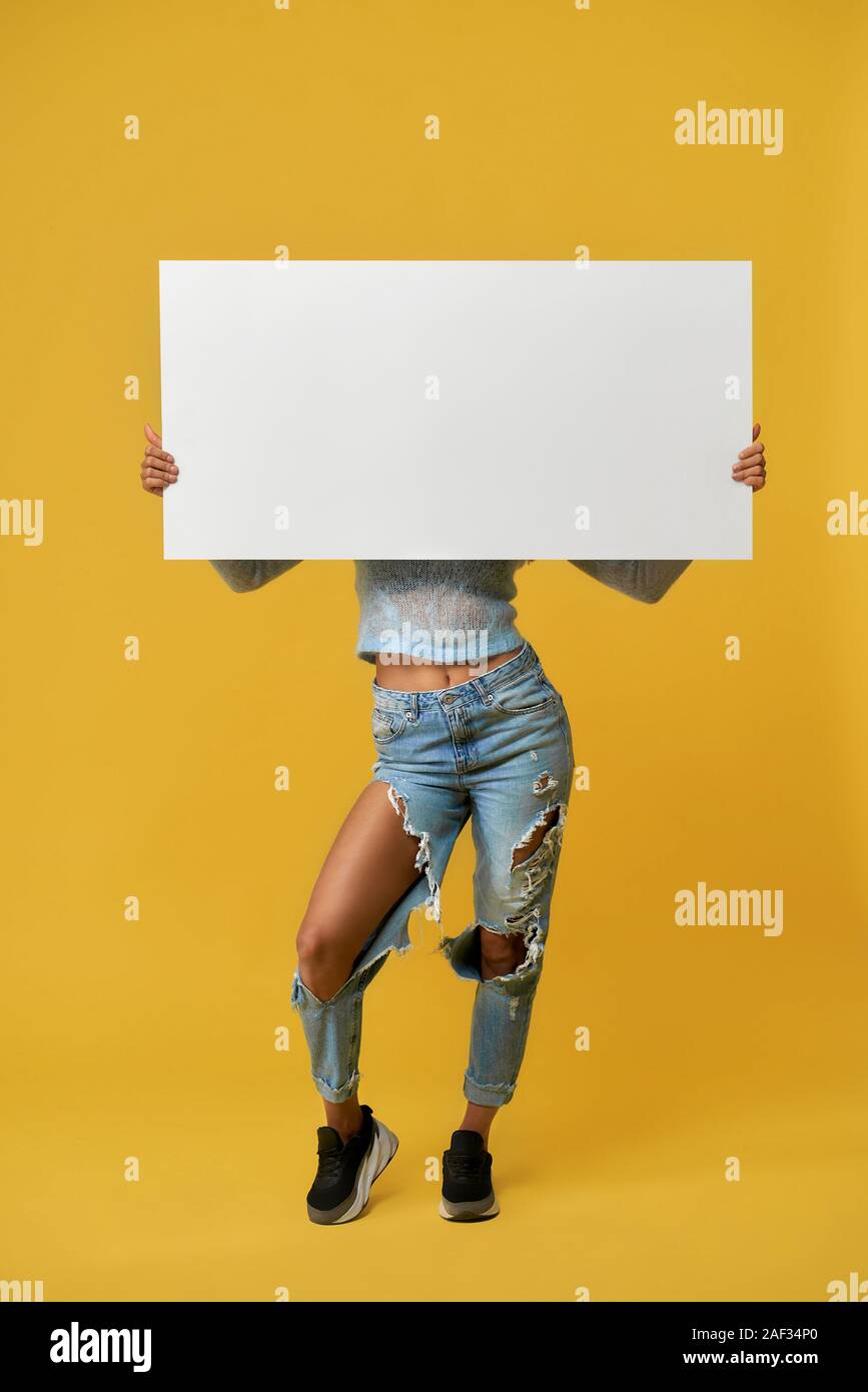 Front view of incognito female in torn jeans holding big banner by hands.  Woman wearing stylish outfit with empty white desk hiding face, standing on  yellow studio background. Concept of advertising Stock