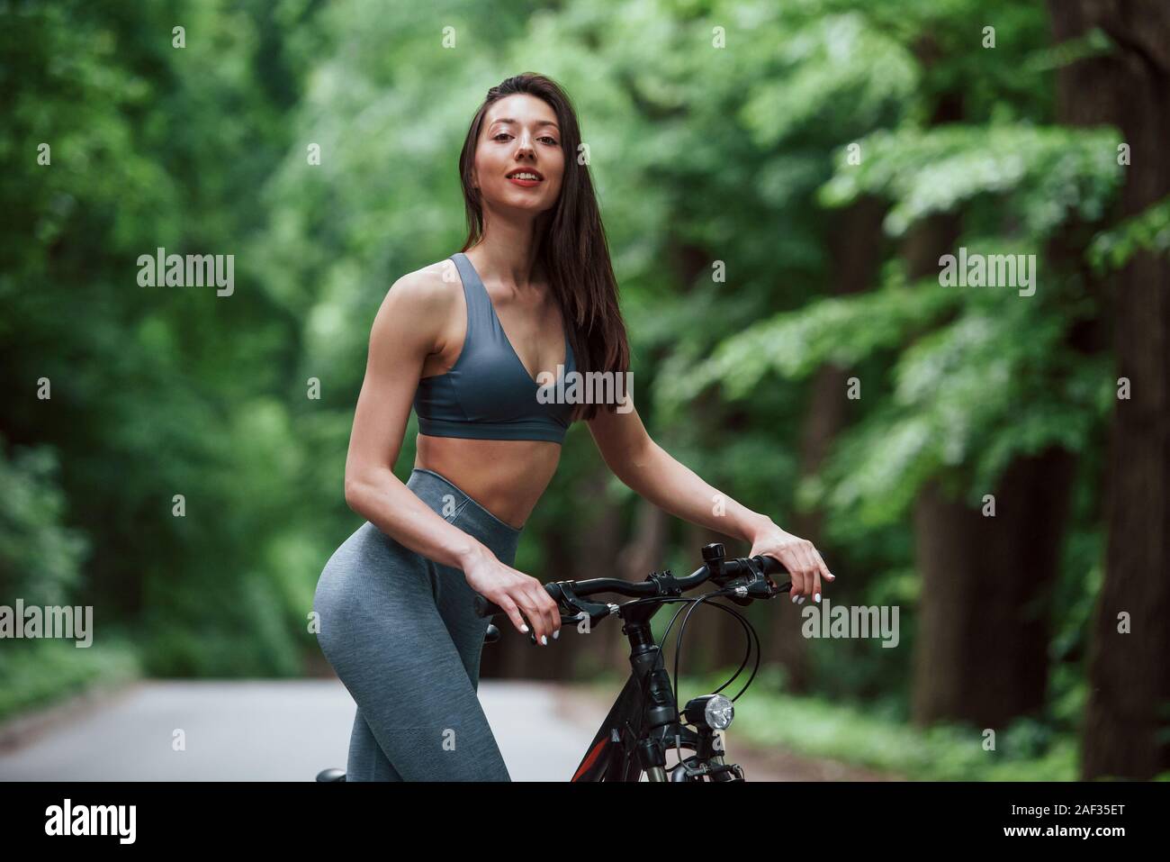 Positive emotions. Female cyclist standing with bike on asphalt road in the forest at daytime Stock Photo