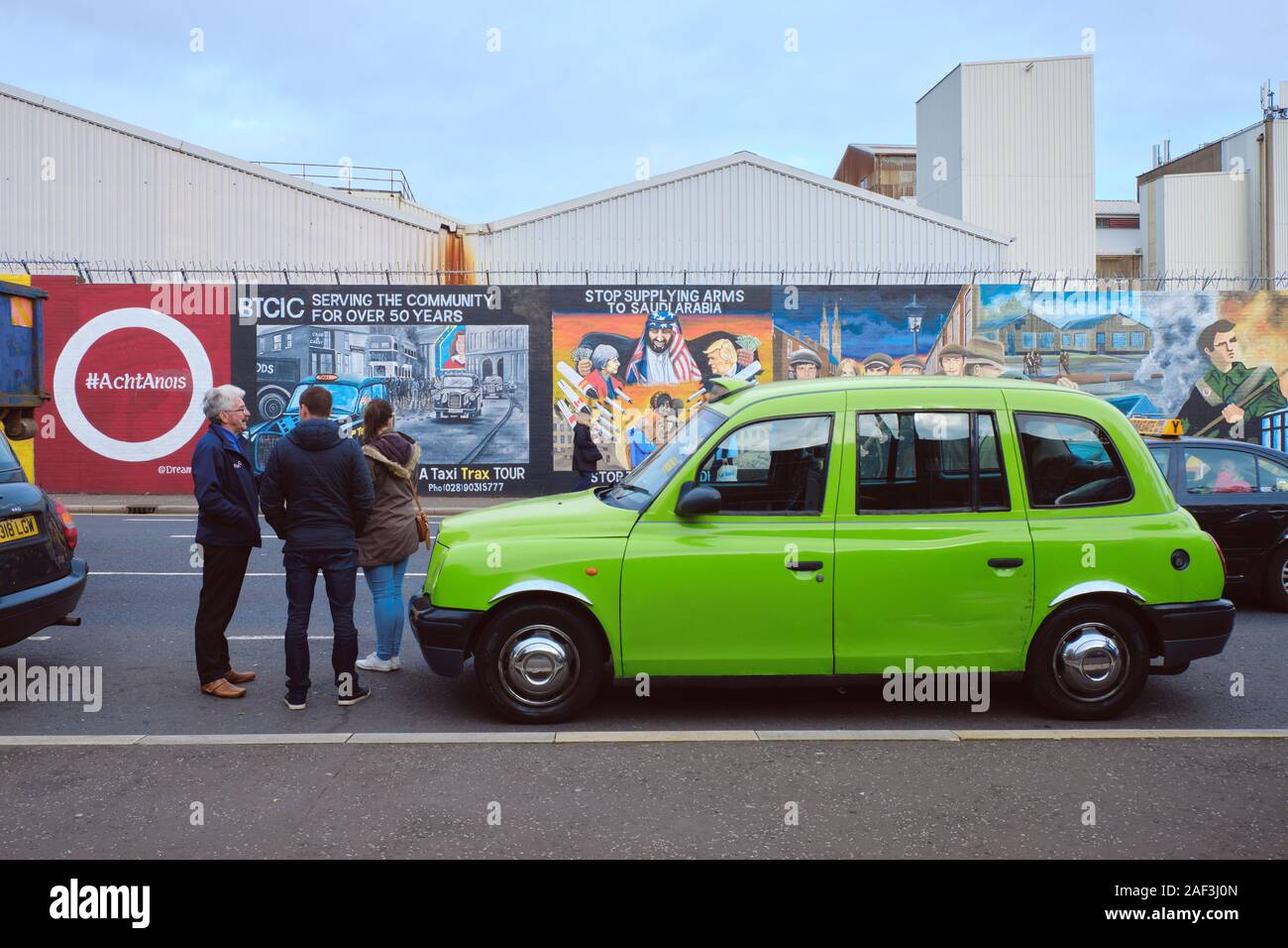 Green traditional cab on a tour of Belfast, with driver giving history of Troubles to two tourists in Front on peace art wall Stock Photo