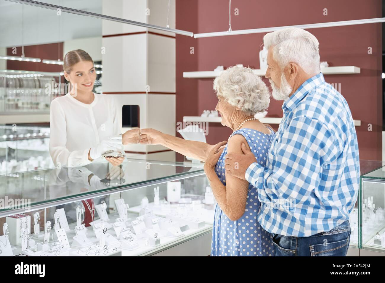 Bearded elder man in checkered shirt embracing his charming wife in blue elegant dress while she trying on new pearl necklace at jewelry store. Caring husband doing luxury present to his beloved woman Stock Photo