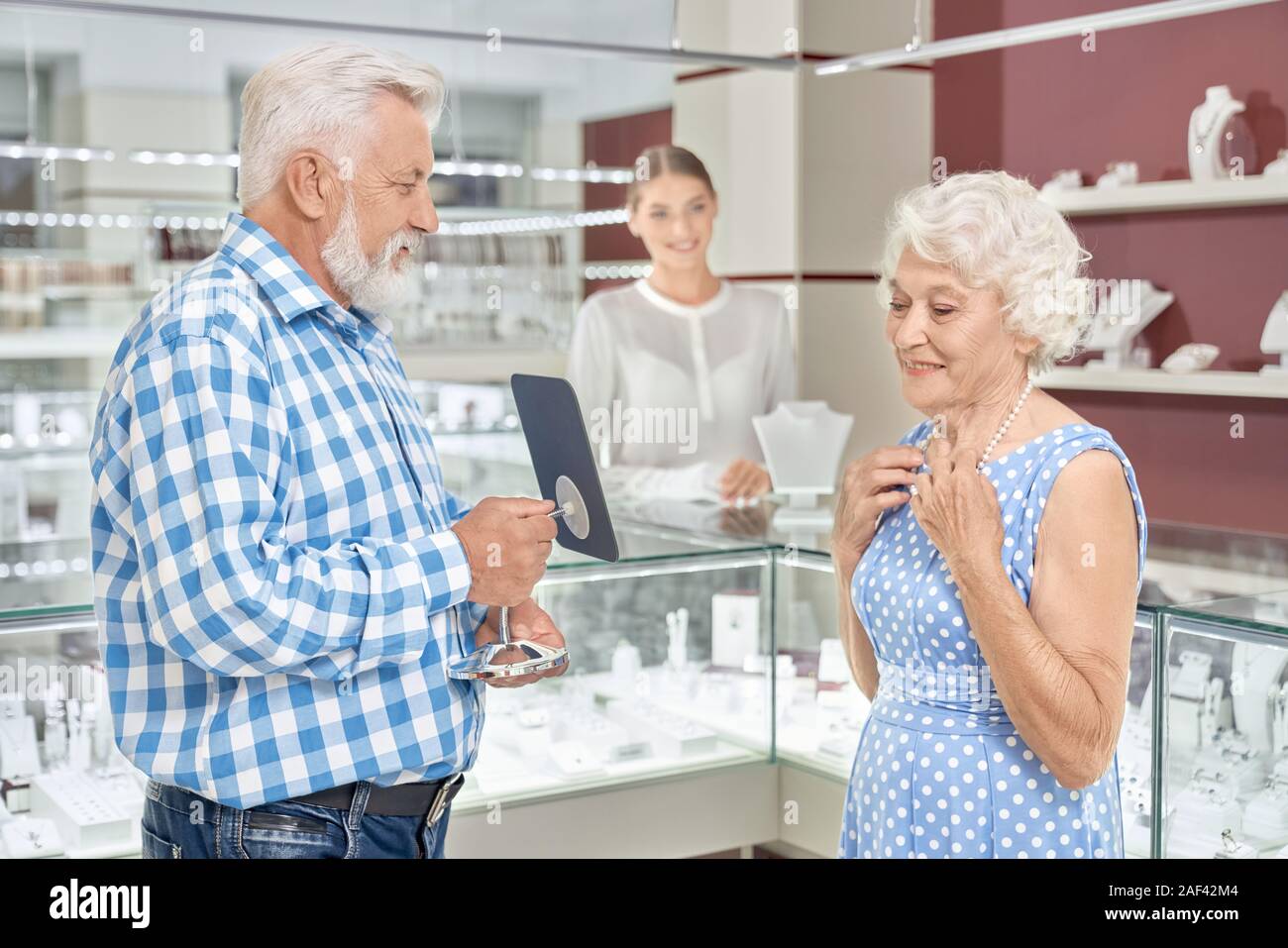 Charming mature woman in elegant dress touching pearl necklace on neck and looking at mirror that holding her bearded senior husband in checkered shirt. Taking pleasure from shopping at jewelry store Stock Photo