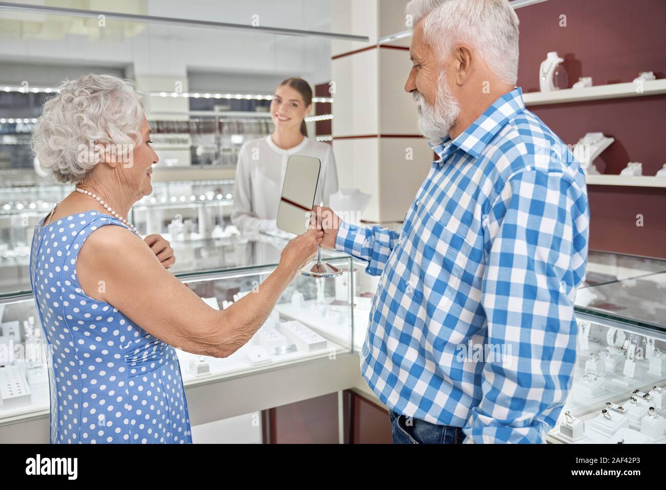 Senior lady with grey hair wearing blue polka dot dress, trying on pearl necklace and looking at mirror that holding her husband in checkered shirt. Happy mature couple buying jewelry in luxury store Stock Photo