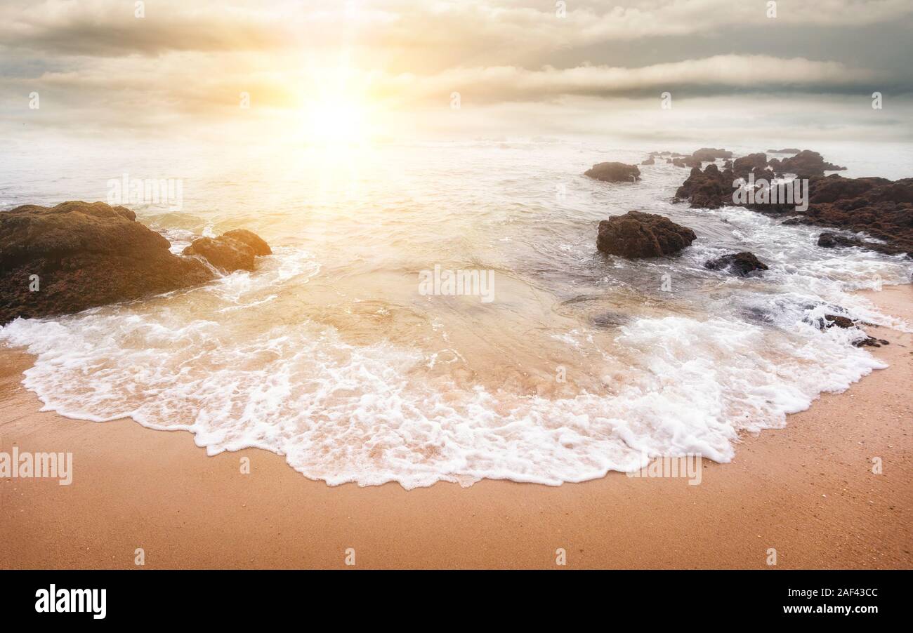 Glorious sunrise scene at the ocean with the sun, clouds and a beautiful little white wave on the sand in the foreground Stock Photo