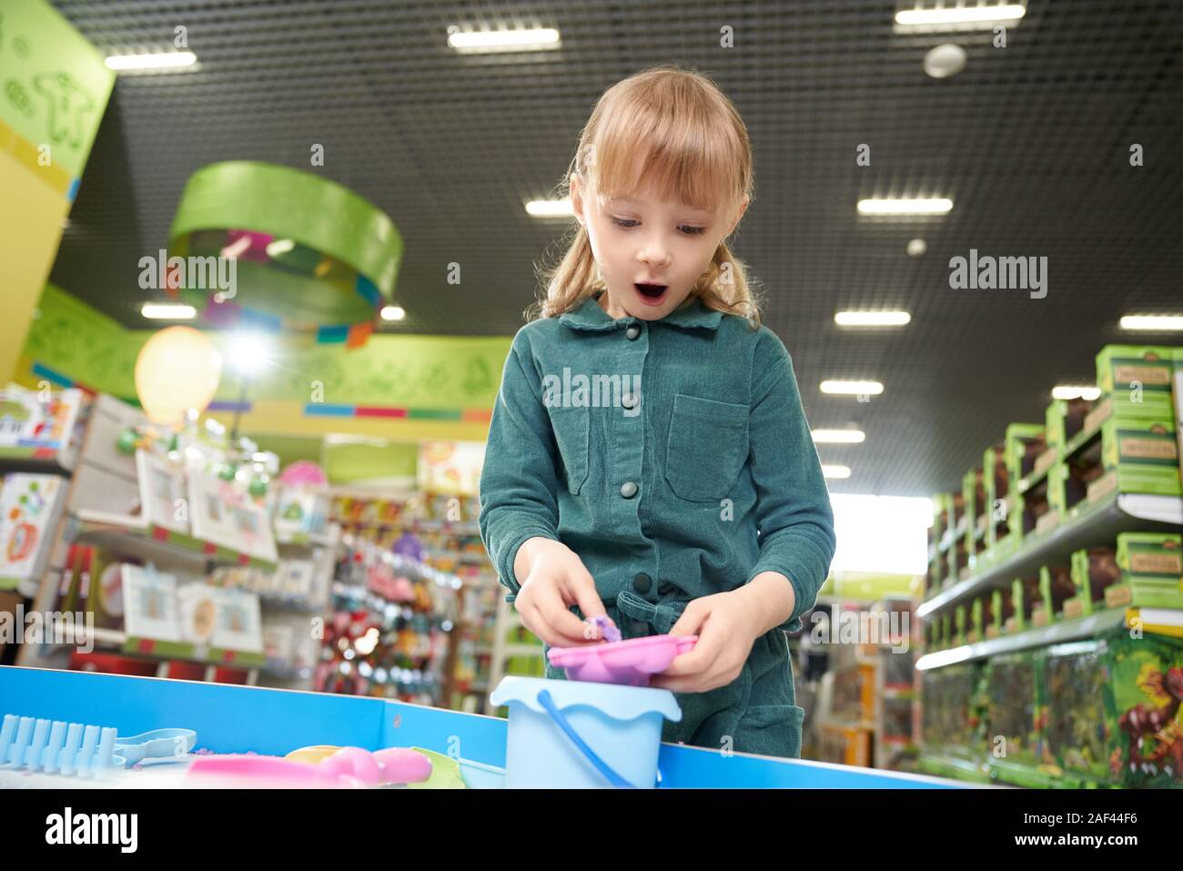 Suprized girl wih open mouth playing with plasticine in big toy store. Little female child enjoying, choosing and buying new cool toys in supermarket. Concept of childhood and joy. Stock Photo
