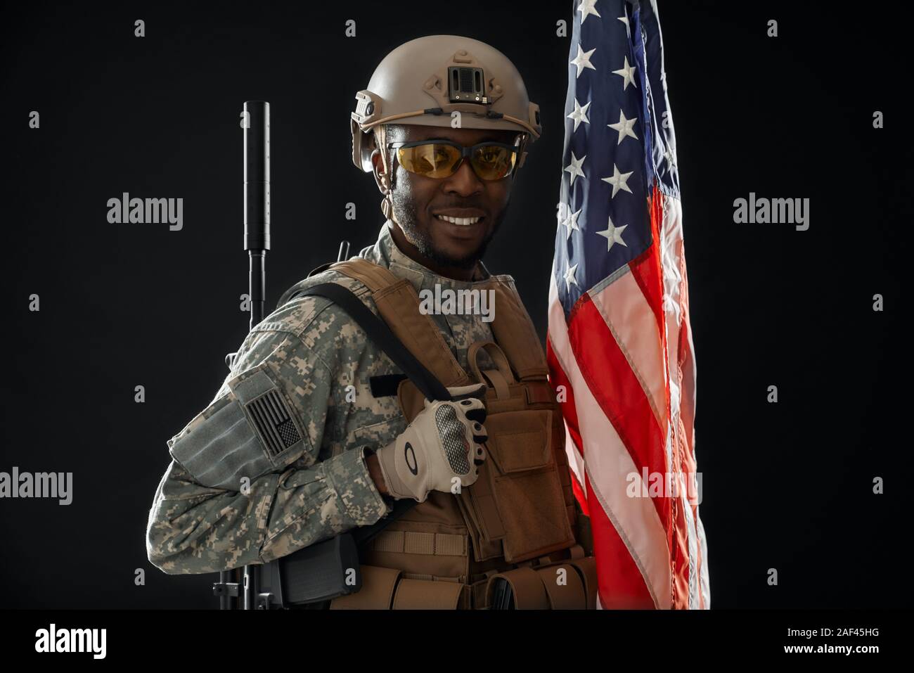 Front view of positivity American soldier wearing army uniform and armour smiling at camera. African man protecting homeland and waiting for instruction, holding weapon on shoulder and national flag. Stock Photo