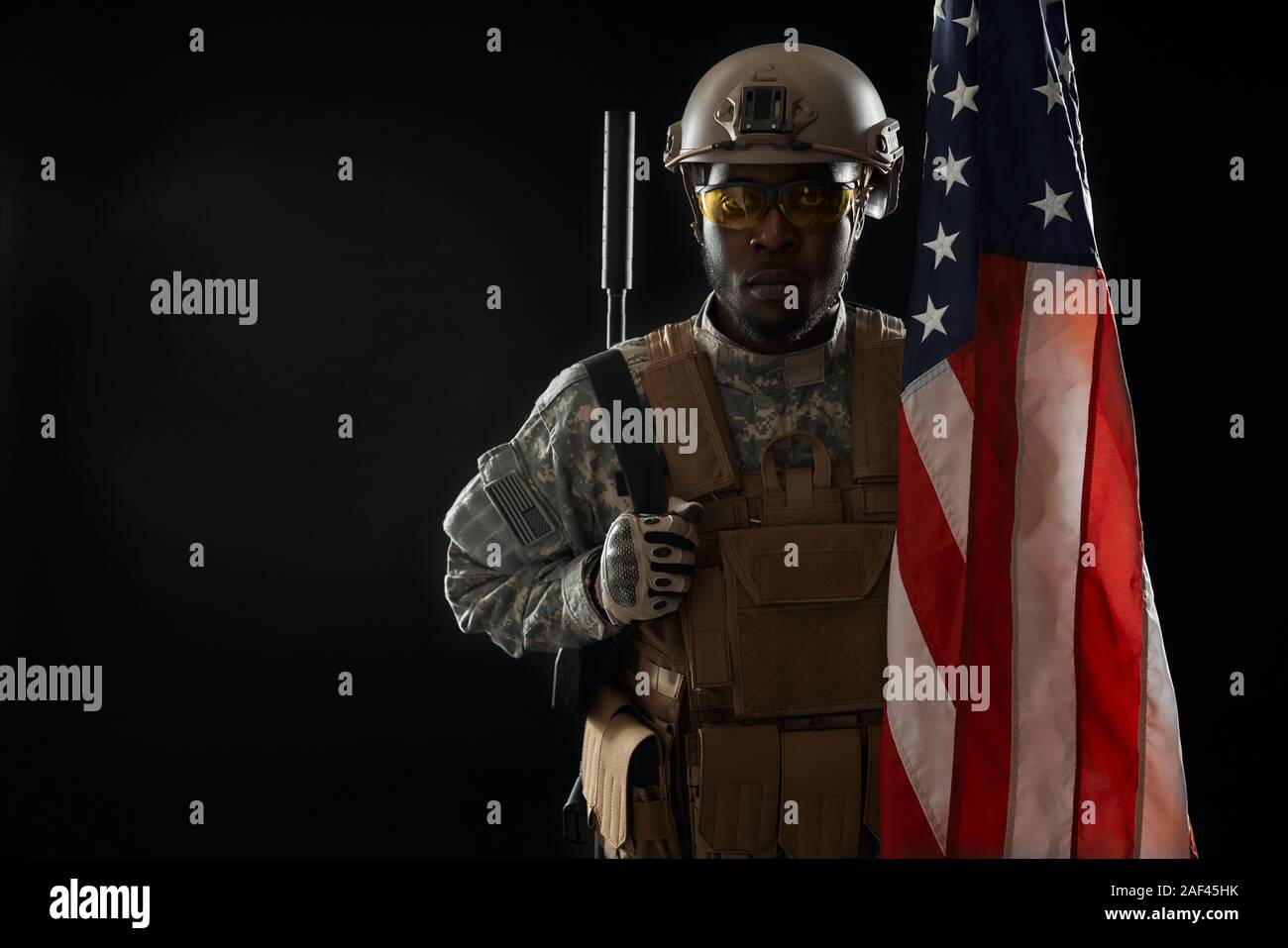 American ranker confidently standing in darkness with weapon and looking at camera. Young African soldier wearing army uniform and armour, posing with American flag. Concept of army. Stock Photo